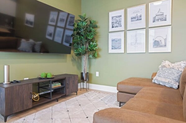 A living room with a dark wood TV stand, a large flat-screen TV, a brown leather sofa, a potted plant, and framed pictures on the wall. - Beltline Lux Loft (Atlanta)