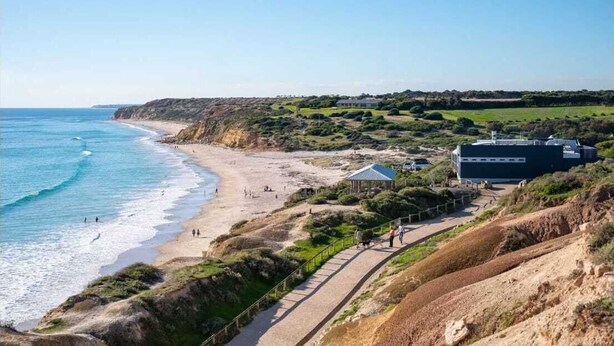 A coastal landscape with a beach, a walking path, and a building with a blue roof.