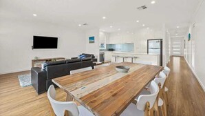 A modern kitchen with white cabinets, a dining area with a wooden table and white chairs, and a living area with a dark sofa and a flat-screen TV mounted on the wall.