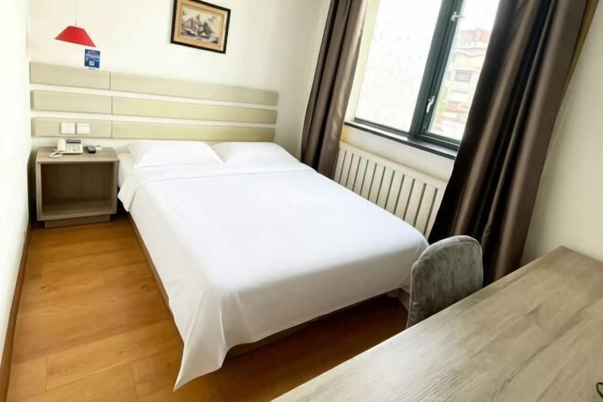A hotel room with a bed, a nightstand with a phone, a desk, a chair, a window with curtains, and a radiator.