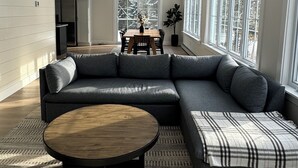 A modern living room with a sectional sofa, a round coffee table, and a dining table with chairs. There are large windows providing natural light and a view of trees outside.