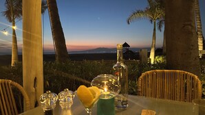A table with a bottle, a candle, and a glass with a heart-shaped garnish, set against a backdrop of palm trees and a sunset view.