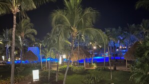 A nighttime resort with palm trees, a pool area, and a thatched-roof structure.
