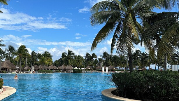 A large, clear swimming pool surrounded by palm trees and thatched-roof structures.
