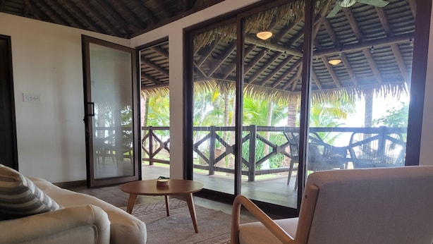 A room with a sofa, a chair, a small table, and a large window with a view of a wooden deck and palm trees. - Stunning Beachfront Home near Cambutal Beach Break (Ave María)