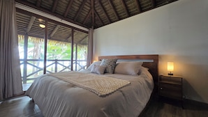 A bedroom with a large bed, a wooden headboard, a bedside table with a lamp, and a view of palm trees through the window. - Stunning Beachfront Home near Cambutal Beach Break (Ave María)