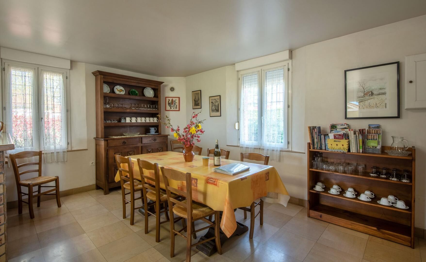 Een eetgedeelte met een tafel voor zes personen, omringd door stoelen. Er is een houten kast met servies en glazen, en een boekenkast met boeken en decoratieve objecten.
