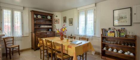 Een eetgedeelte met een tafel voor zes personen, omringd door stoelen. Er is een houten kast met servies en glazen, en een boekenkast met boeken en decoratieve objecten.