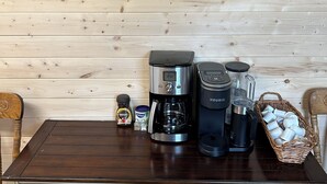 Una zona de cocina con una cafetera, una licuadora y un hervidor sobre una mesa de madera. Hay un letrero enmarcado en la pared y un cesto de tazas sobre la mesa.