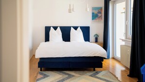 A bedroom with a large bed, a blue headboard, white pillows, a patterned rug, a window with curtains, and a wall-mounted light fixture.
