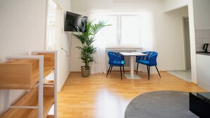 A modern living room with a wooden floor, a dining table with blue chairs, a TV mounted on the wall, and a potted plant.