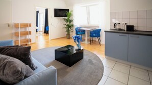 A modern living room with a grey sofa, a black coffee table, and a blue chair. There is a kitchen area with a blue chair and a white tiled backsplash.