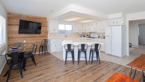 Una cocina moderna con armarios blancos, una isla central con taburetes, un televisor de pantalla plana montado en una pared de madera, y un comedor con mesa y sillas de madera.