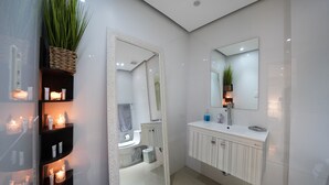 Un baño moderno con azulejos blancos, un espejo grande, un lavabo blanco con encimera y repisas con velas.