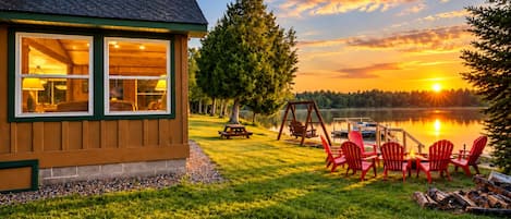 A cabin with a view of a lake at sunset, outdoor seating, and a swing set.