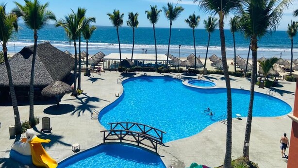 A pool area with a bridge, palm trees, and a beach view.