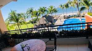 A balcony with a table and chairs, overlooking a swimming pool and beach with palm trees.
