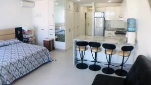 A compact living space with a kitchenette, a bed with patterned bedding, a ceiling fan, and a water dispenser. - Aria Ocean Beachfront Condos (Nuevo Nayarit)