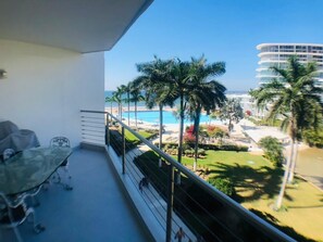 Un balcón con una mesa y sillas de vidrio, con vistas a una piscina y palmeras.