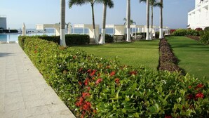 A well-maintained garden with palm trees, a swimming pool, and a white building with multiple balconies. - Aria Ocean Beachfront Condos (Nuevo Nayarit)