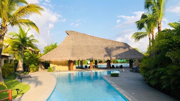 Una piscina con una estructura de techo de paja al fondo, rodeada de palmeras y un cielo despejado.