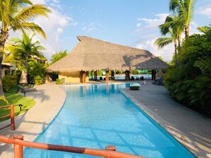 A swimming pool with a thatched-roof structure in the background, surrounded by palm trees and a clear sky. - El Tigre Isla Palmares (Nuevo Nayarit)
