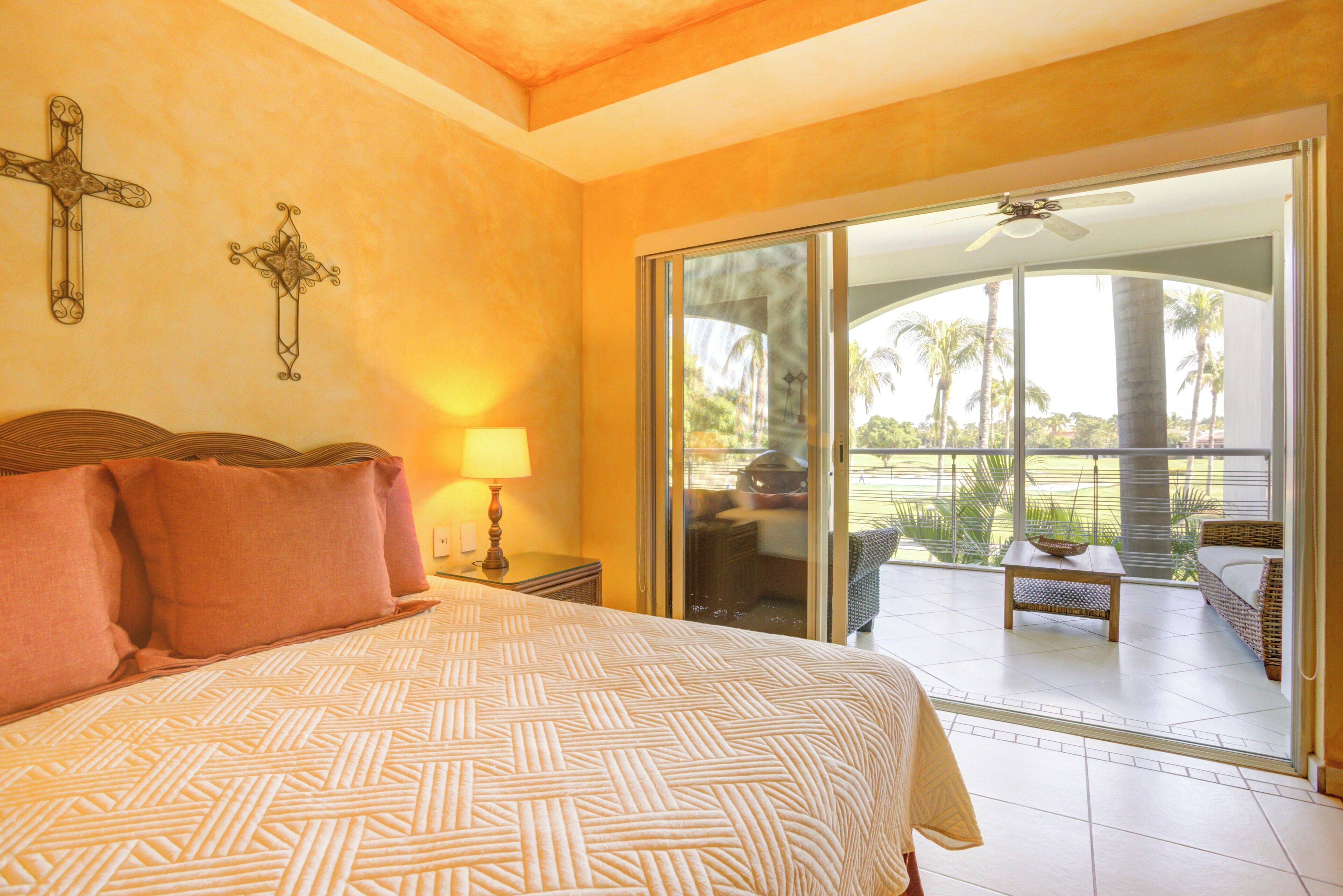 A bedroom with a large bed, a nightstand with a lamp, and a sliding glass door leading to a balcony with outdoor furniture.