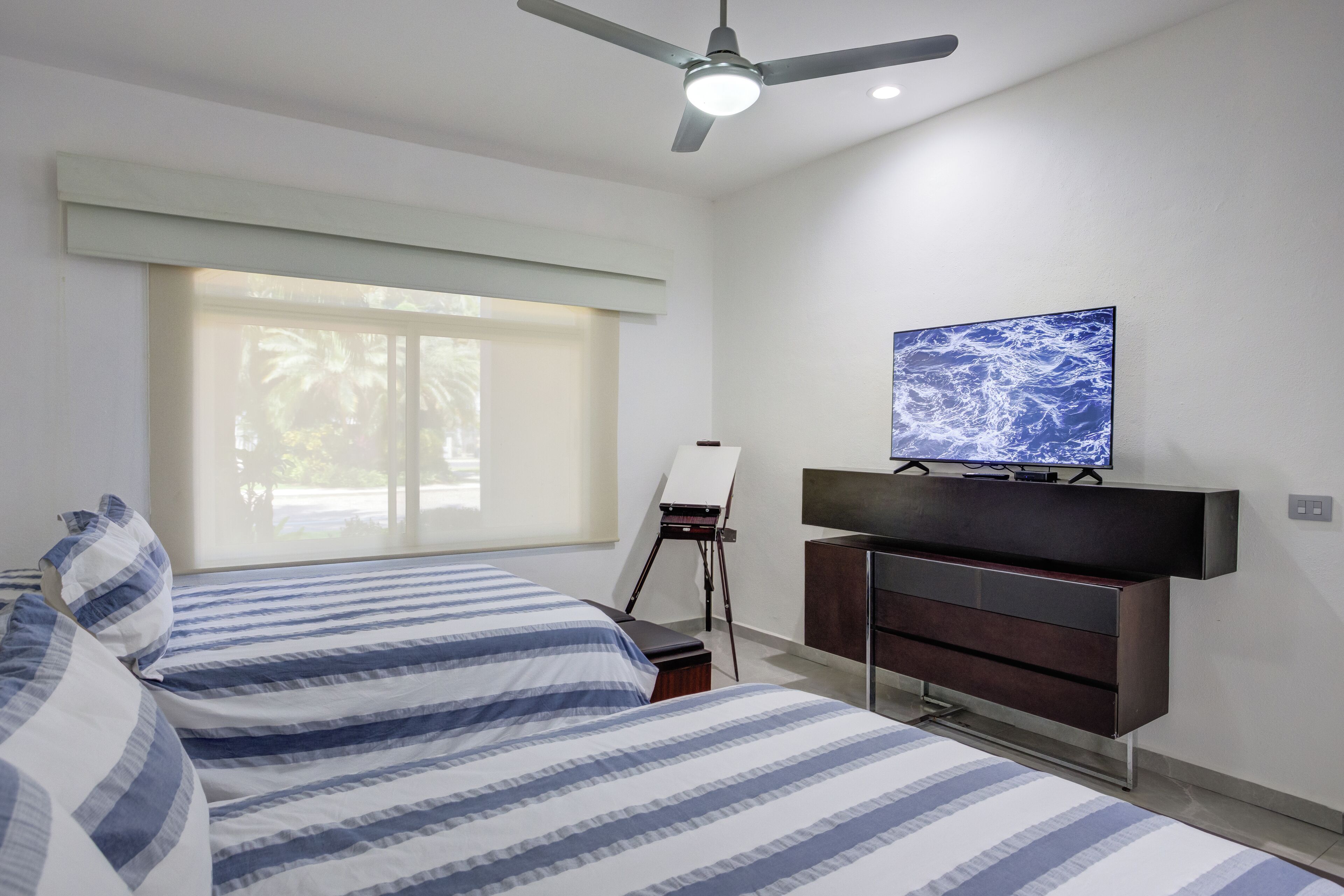 A bedroom with a bed, a dresser, a painting on a easel, and a television on the wall.