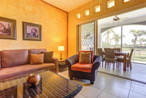 A living room with a sofa, armchair, and coffee table, a dining area with chairs and a table, and a large sliding glass door leading to a balcony with palm trees. - El Tigre Green Bay I Condos (Nuevo Nayarit)
