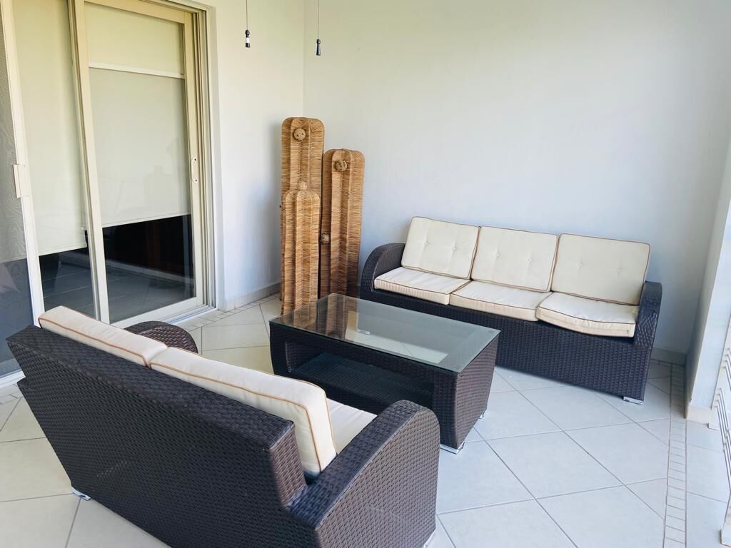 Un espace extérieur moderne aménagé avec un canapé et deux fauteuils, une table basse avec un plateau en verre et une porte-fenêtre coulissante donnant sur un balcon.