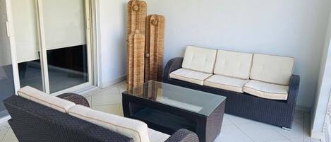 Un espace extérieur moderne aménagé avec un canapé et deux fauteuils, une table basse avec un plateau en verre et une porte-fenêtre coulissante donnant sur un balcon.