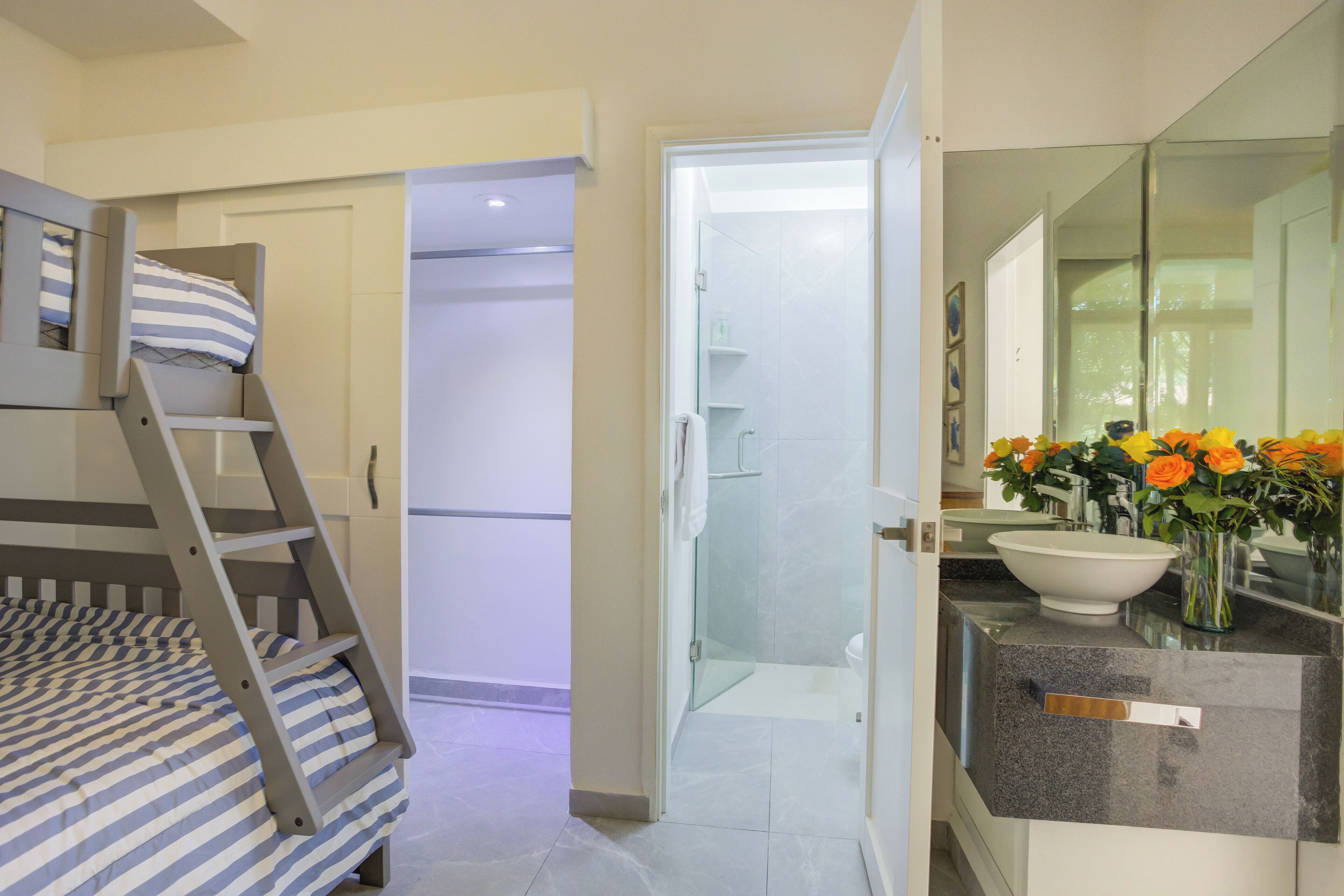 A bedroom with a bunk bed, a bathroom with a glass shower, and a sink with a vase of flowers.