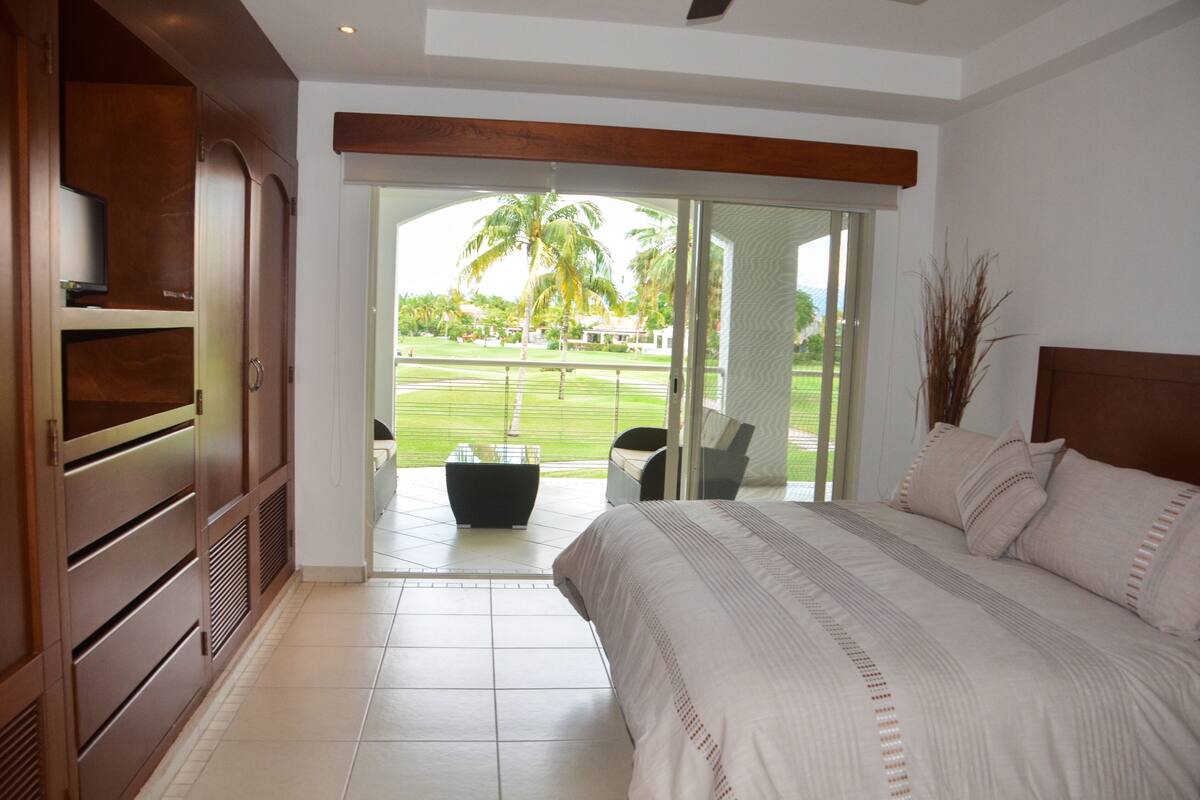 A bedroom with a large bed, a TV, and a sliding glass door leading to a balcony with a view of a golf course.
