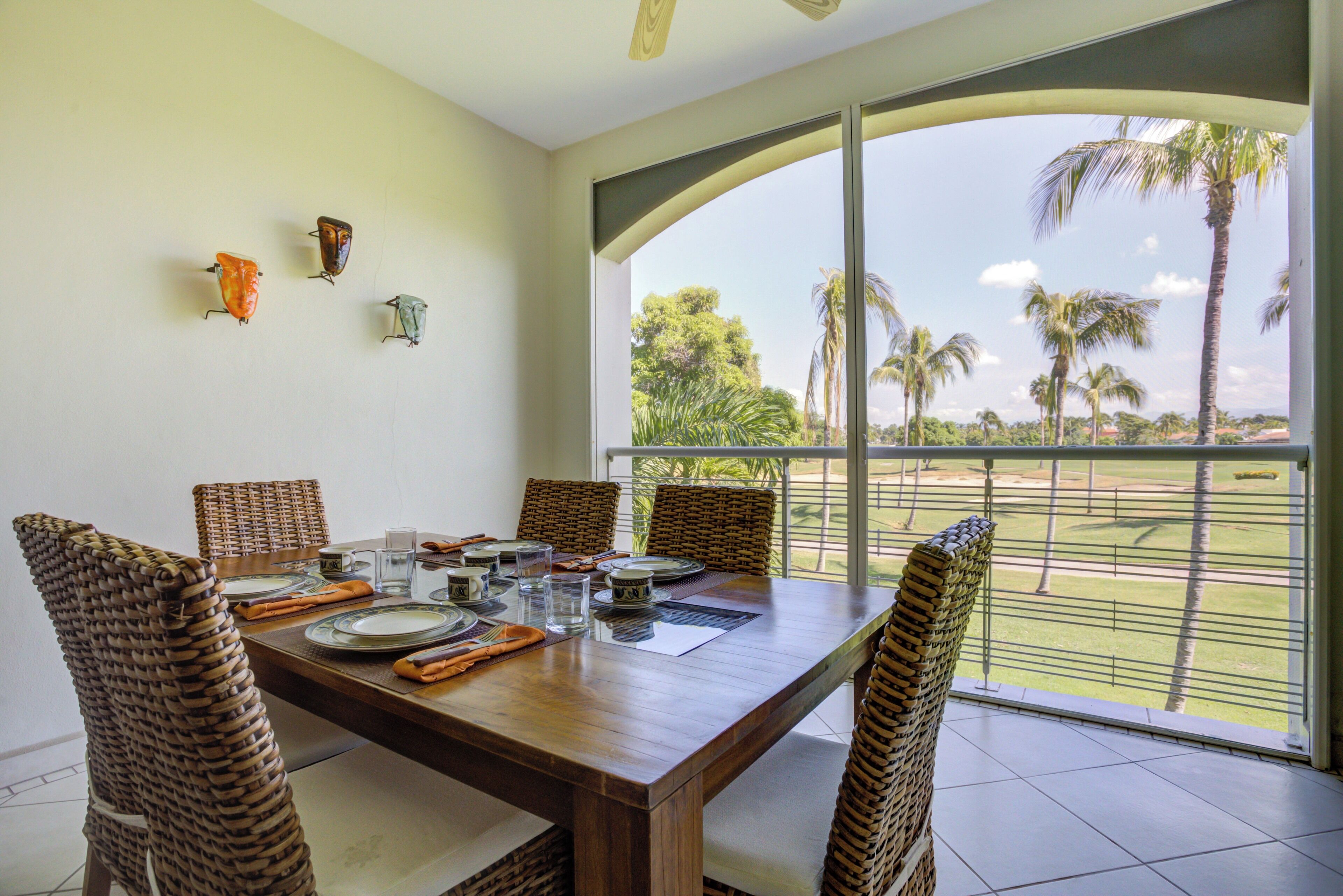 A dining area with a wooden table set for four, wicker chairs, and a view of palm trees and greenery through large windows.