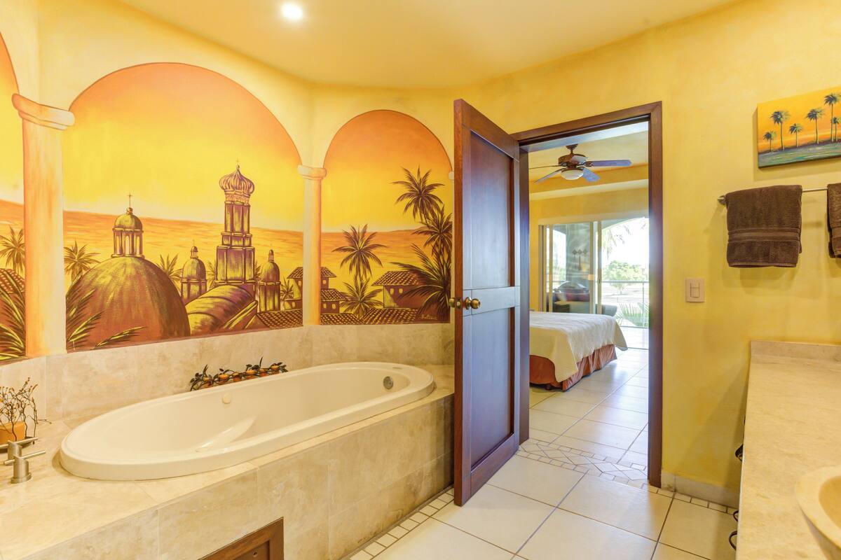 A bathroom with a large bathtub, a shower, and a toilet. There is a mural on the wall depicting a sunset scene.