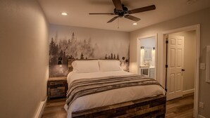 A bedroom with a bed, a ceiling fan, a nightstand, a wall-mounted lamp, and a door leading to a bathroom.