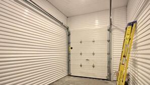 Un couloir étroit, avec des portes enroulables fermées de chaque côté, une échelle jaune appuyée contre l’un des murs et un sol en béton.