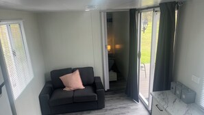 Una habitación moderna con un sofá gris oscuro, una mesita de noche con una lámpara y vistas a un jardín por una puerta corredera.