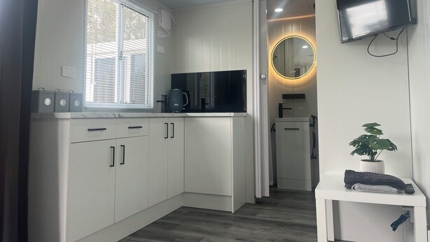 Una cocina moderna con armarios blancos, encimera negra, una ventana con persianas, televisión integrada, un espejo redondo y una pequeña mesa con una planta y una toalla.