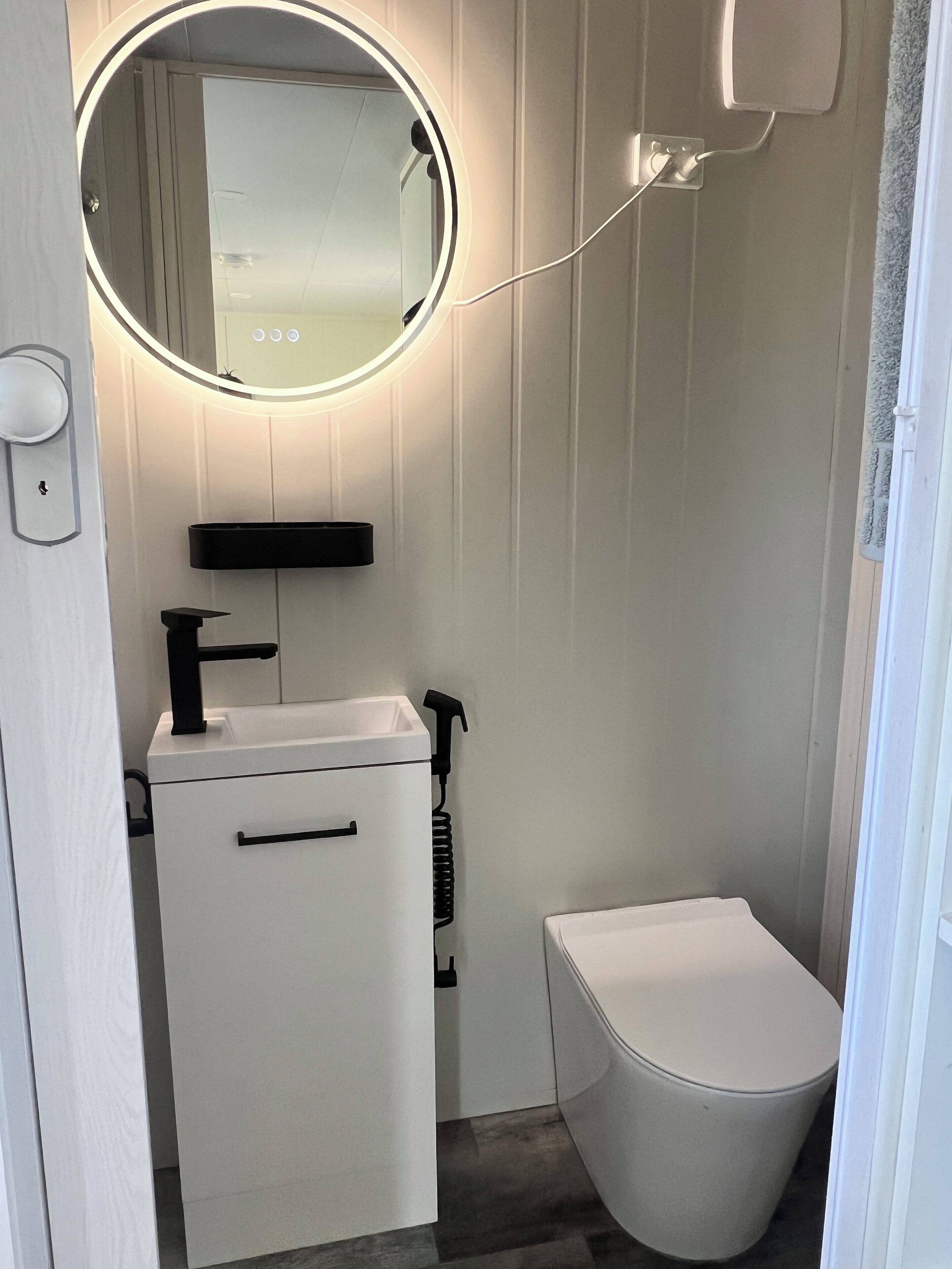 Een kleine, moderne badkamer met een witte wastafel, een spiegel met verlichting, een toilet en een wandplankje.