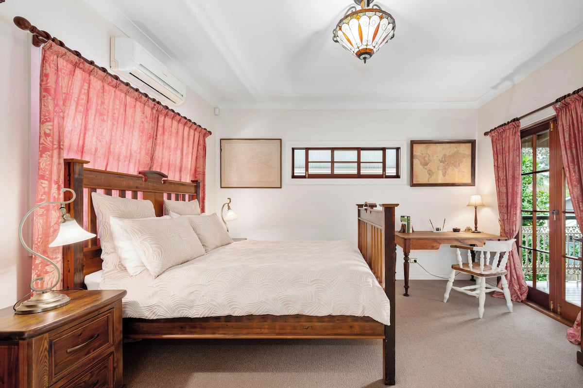 A bedroom with a wooden bed, a desk, and a chair. There is a window with curtains, a map on the wall, and a lamp on the nightstand.