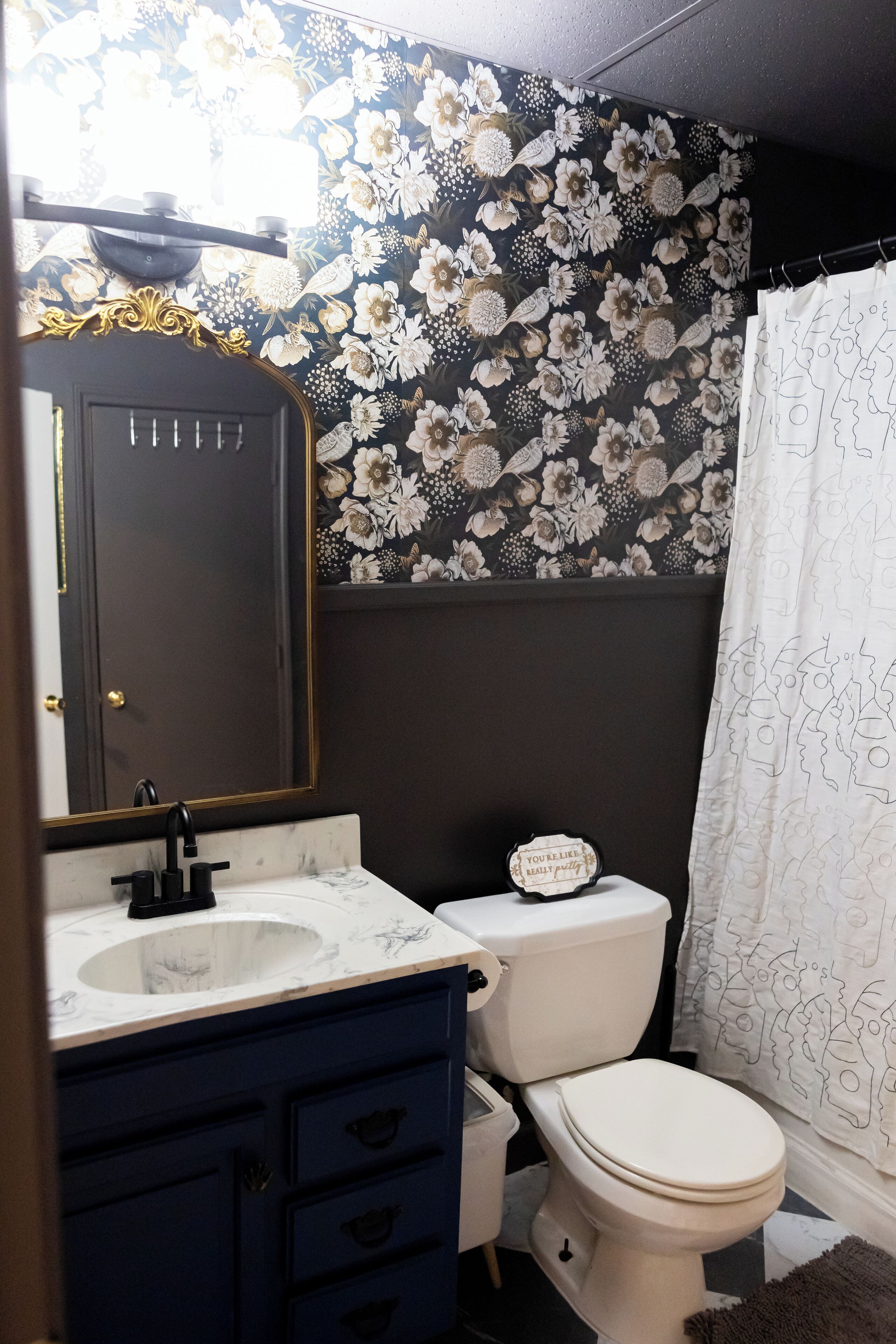 Een badkamer met een witte wasbak, een donkerblauwe opbergkast, een wit toilet en een douchegordijn met een bloemenprint.
