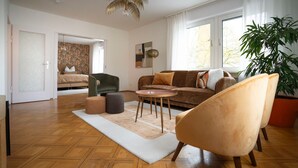 Ein geräumiges Wohnzimmer mit Fischgrätparkett, einem beigefarbenen Sofa, einem runden Couchtisch und einer großen Küstpflanze.