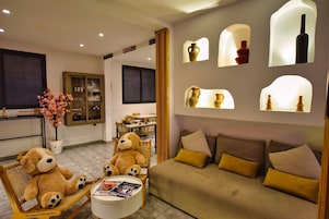 Un salón acogedor con un sofá, dos sillones y una mesita baja con revistas. Se observan estanterías decorativas con jarrones y una planta en maceta.