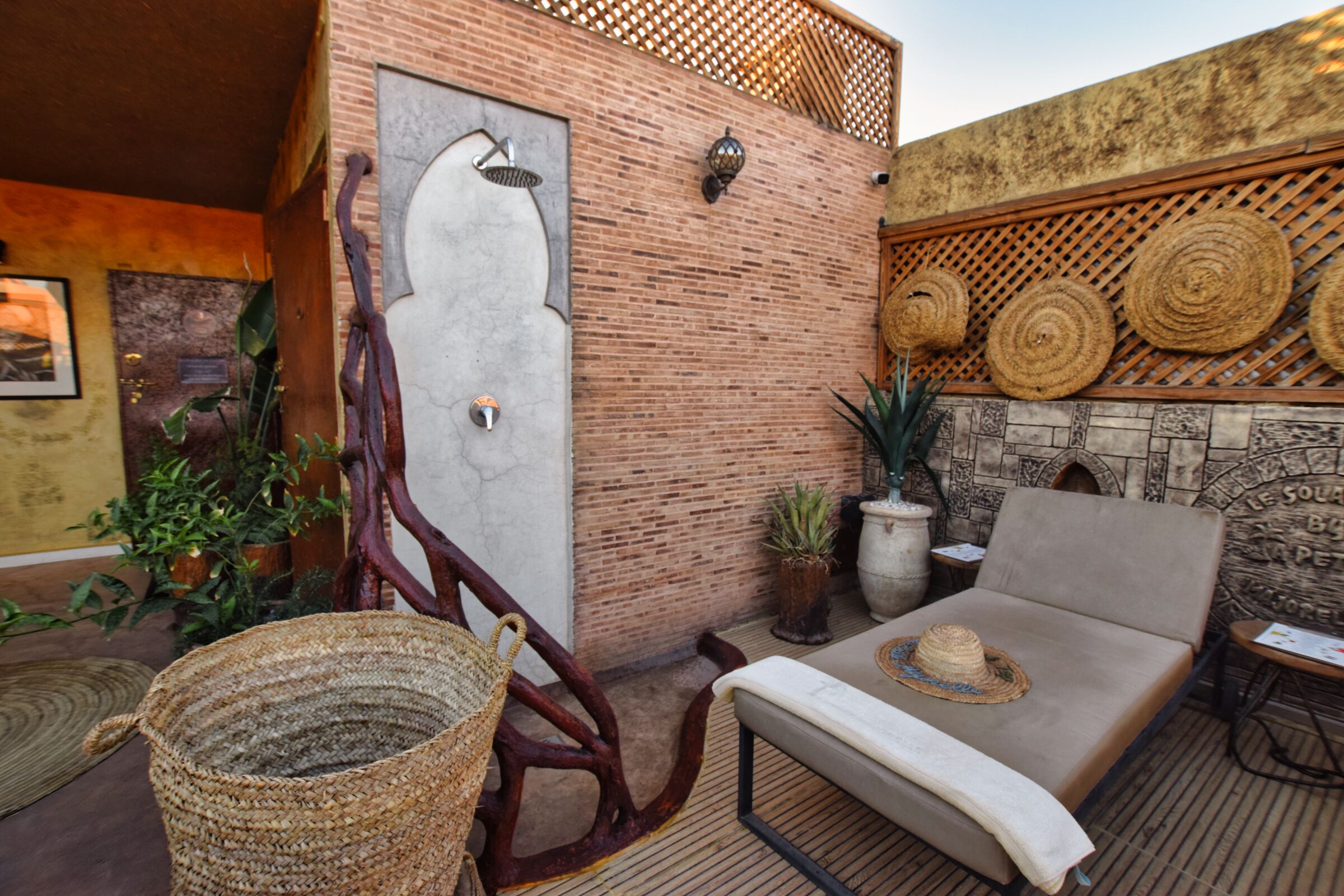 Un espacio exterior acogedor con una silla acolchada, una cesta tejida y un cabezal de ducha empotrado en la pared.