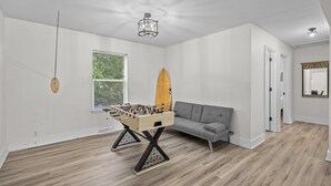 Une pièce avec un plancher en bois, un canapé gris, une table de baby-foot, une planche de surf et un lustre.