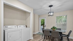Une buanderie équipée d’un lave-linge et d’un sèche-linge, un coin repas avec une table et des chaises, et une fenêtre avec des stores.