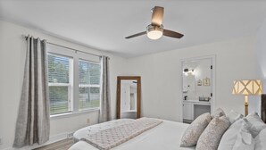Une chambre à coucher avec un lit, un ventilateur de plafond, une fenêtre avec des rideaux, un miroir et une lampe de chevet.