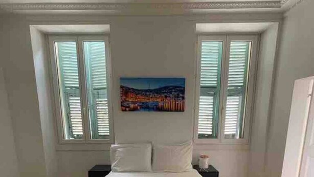A neatly made bed with two folded towels, a painting of a cityscape on the wall, and a window with shutters. - Delightful Haven, Center of Villefranche  (Villefranche-sur-Mer)