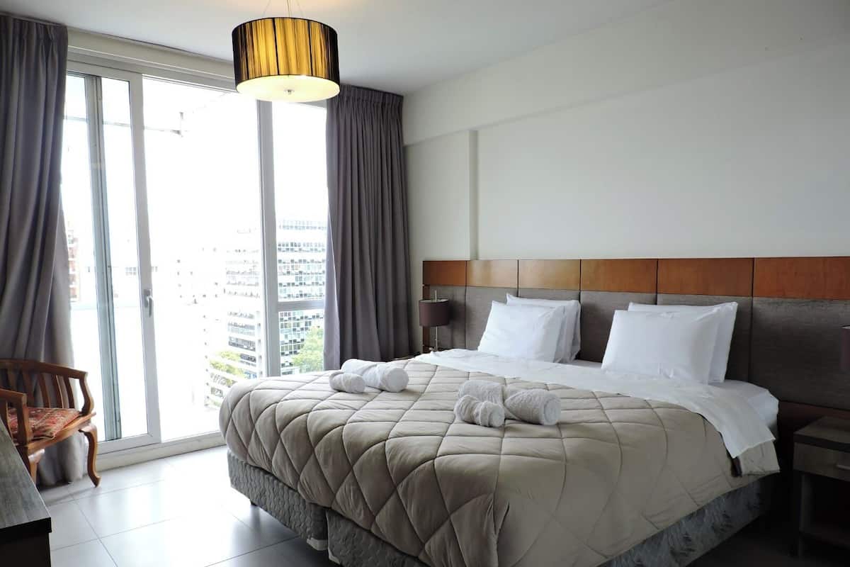 A hotel room with a large bed, a wooden headboard, a bedside table, a chair, and a view of the city through the window.